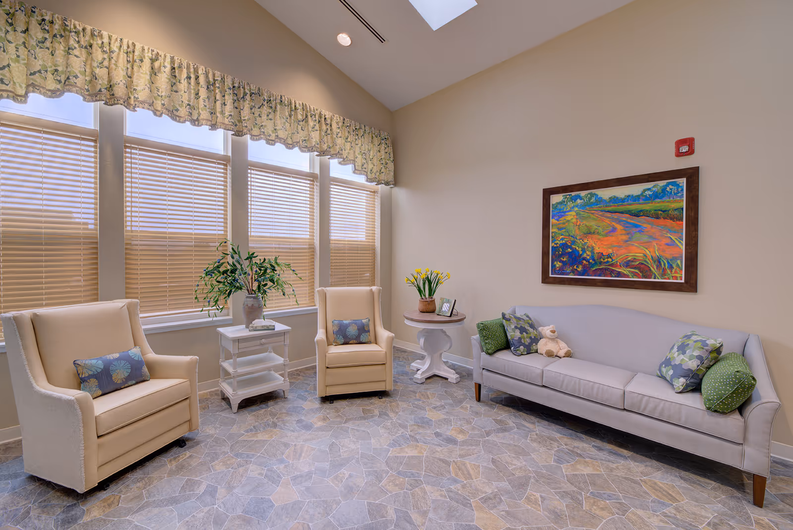 A cozy sitting area with two beige armchairs and a light gray sofa adorned with patterned and green pillows. There are two small tables, one with a vase of green plants and the other with a pot of yellow flowers and a framed photo. A colorful landscape painting hangs on the beige wall above the sofa. The floor has a stone tile pattern, and large windows with beige blinds and floral valances let in natural light.