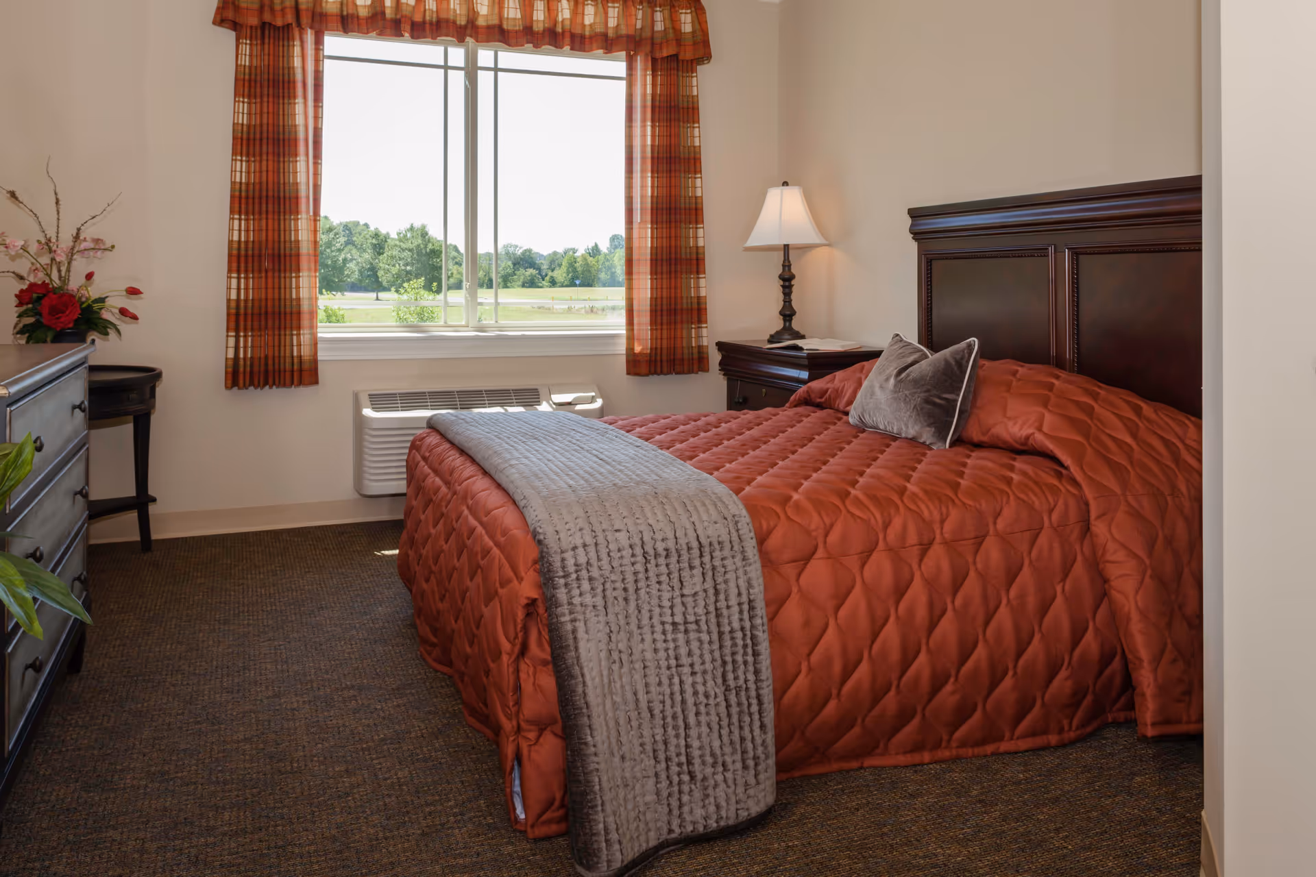 A cozy bedroom with a large window showing a green outdoor view. The bed has a dark wooden headboard and is covered with a rust-colored quilt and a gray throw blanket. There is a nightstand with a lamp and an open book next to the bed, a dresser with flowers on top, and plaid curtains framing the window.