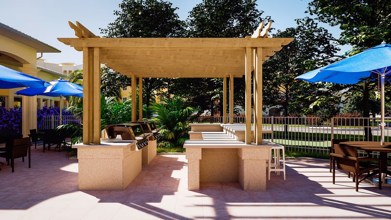 Outdoor patio area with a wooden pergola covering built-in grills and countertops. There are blue umbrellas shading tables and chairs on either side, surrounded by greenery and trees in the background.