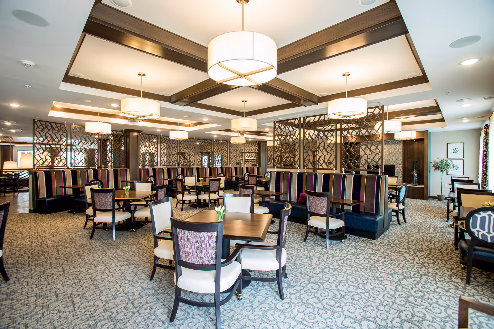 A spacious and well-lit dining room with multiple tables and chairs arranged neatly. The room features patterned carpet flooring, decorative wooden partitions, and ceiling lights with round fabric shades. The seating includes both upholstered chairs and booth-style benches with striped fabric. There are some small flower arrangements on the tables and framed artwork on the walls.