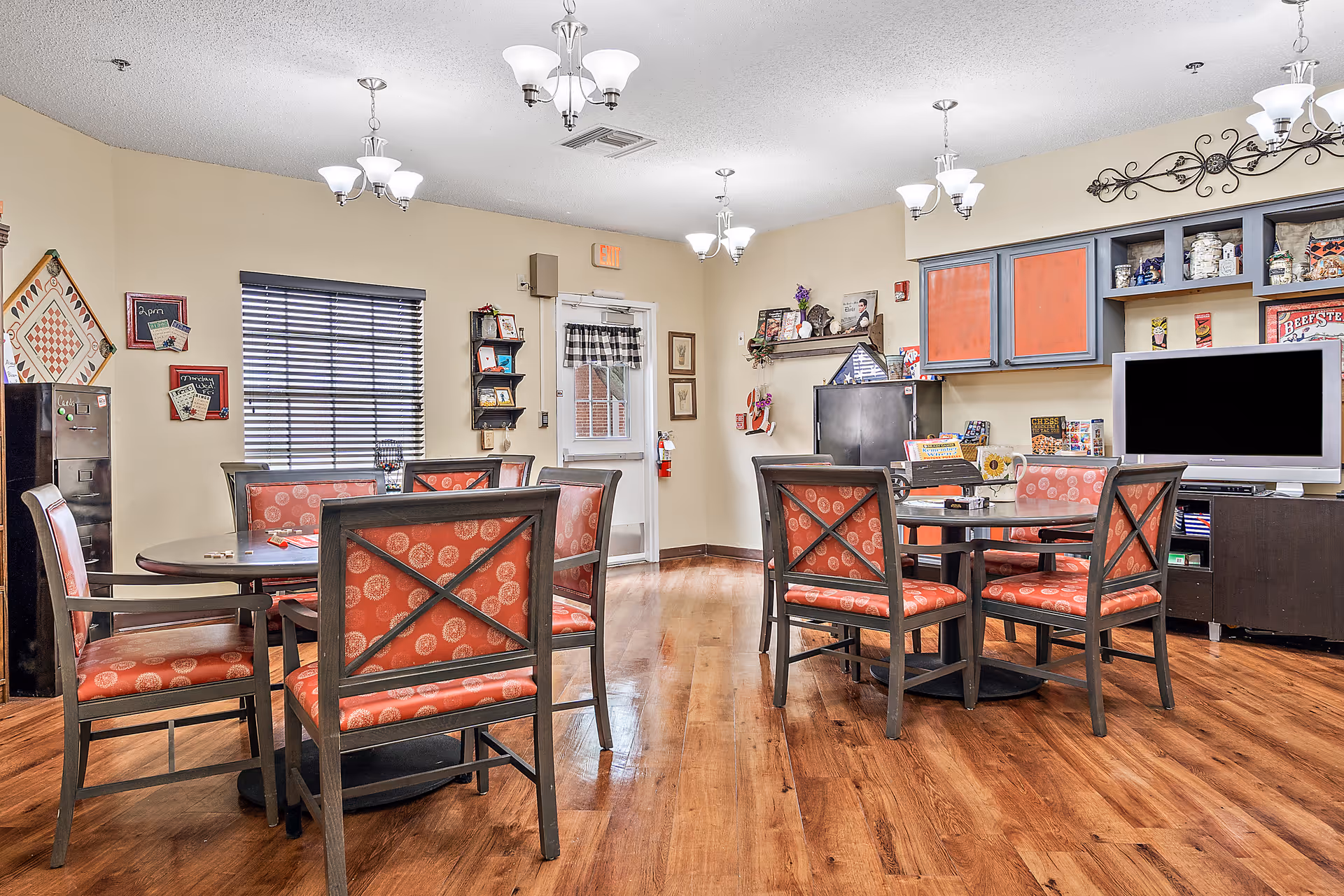 A cozy common area with two round tables surrounded by chairs with red patterned cushions. The room has wooden flooring, beige walls, and multiple ceiling light fixtures. There is a TV on a stand, shelves with various items, a window with blinds, and a door with a small curtain. The space appears warm and inviting, suitable for socializing or activities.