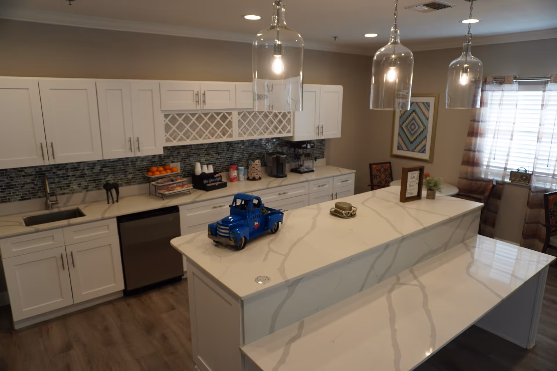 A modern kitchen with white cabinets, a marble countertop island, and three pendant lights hanging from the ceiling. The backsplash is made of small blue and gray tiles. On the countertop, there is a small blue toy truck, a decorative bowl, and a framed sign. In the background, there is a window with striped curtains and a small round table with chairs.