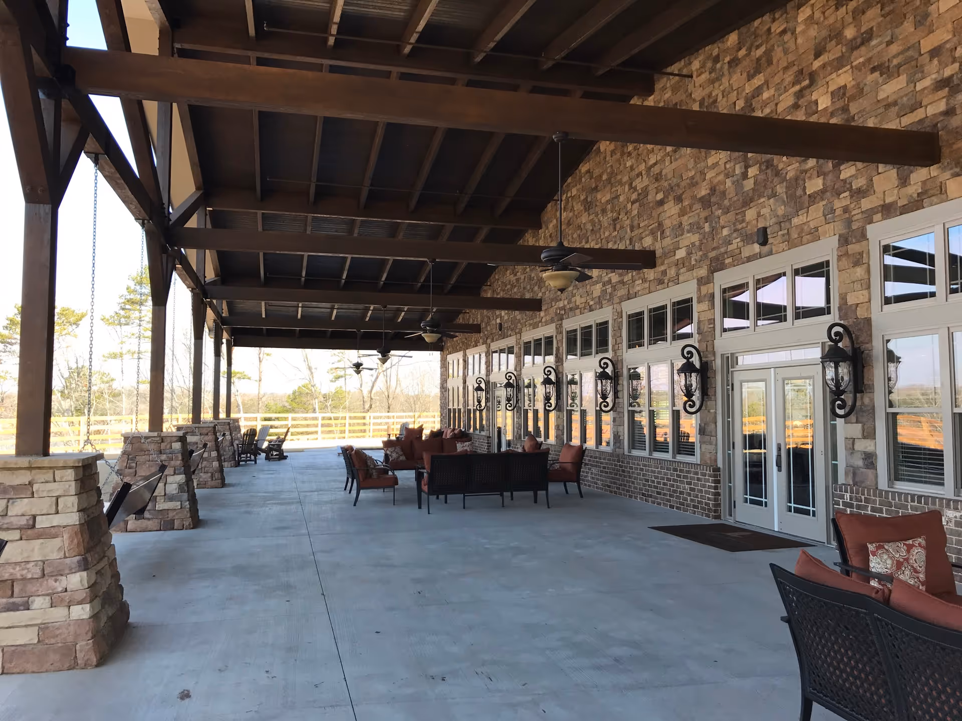 Covered outdoor patio area with stone pillars and wooden beams supporting the roof. The patio has multiple ceiling fans, wall-mounted lantern-style lights, and several seating arrangements with cushioned chairs and sofas. Large windows and double doors line the brick wall of the building, and trees are visible in the background.