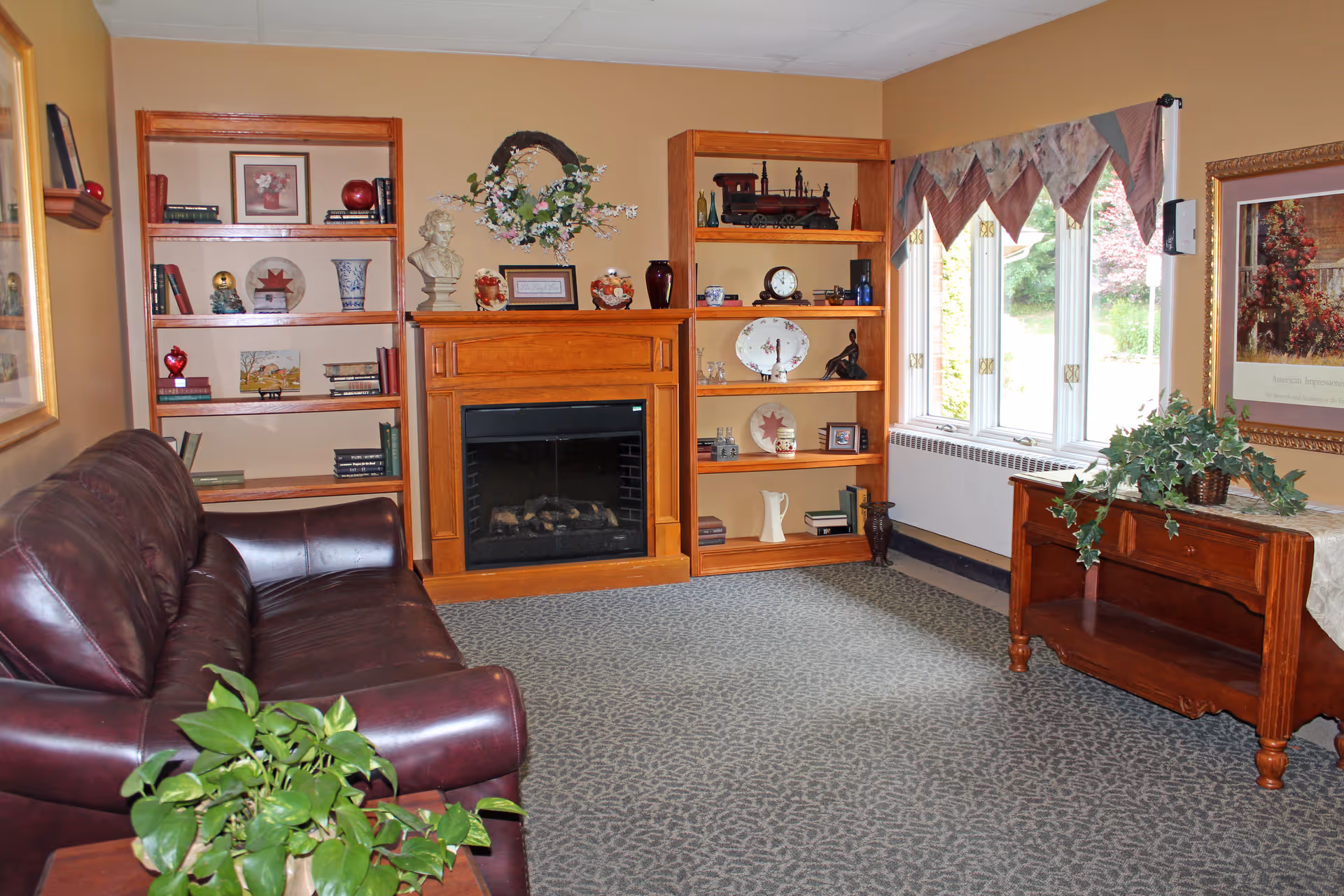 A cozy living room with a dark brown leather sofa on the left, two wooden bookshelves flanking a wooden fireplace mantel decorated with a floral wreath and various ornaments. To the right, there is a window with patterned valances letting in natural light, and a wooden side table with a green leafy plant and a framed picture on the wall above it. The room has beige walls and a patterned carpeted floor.