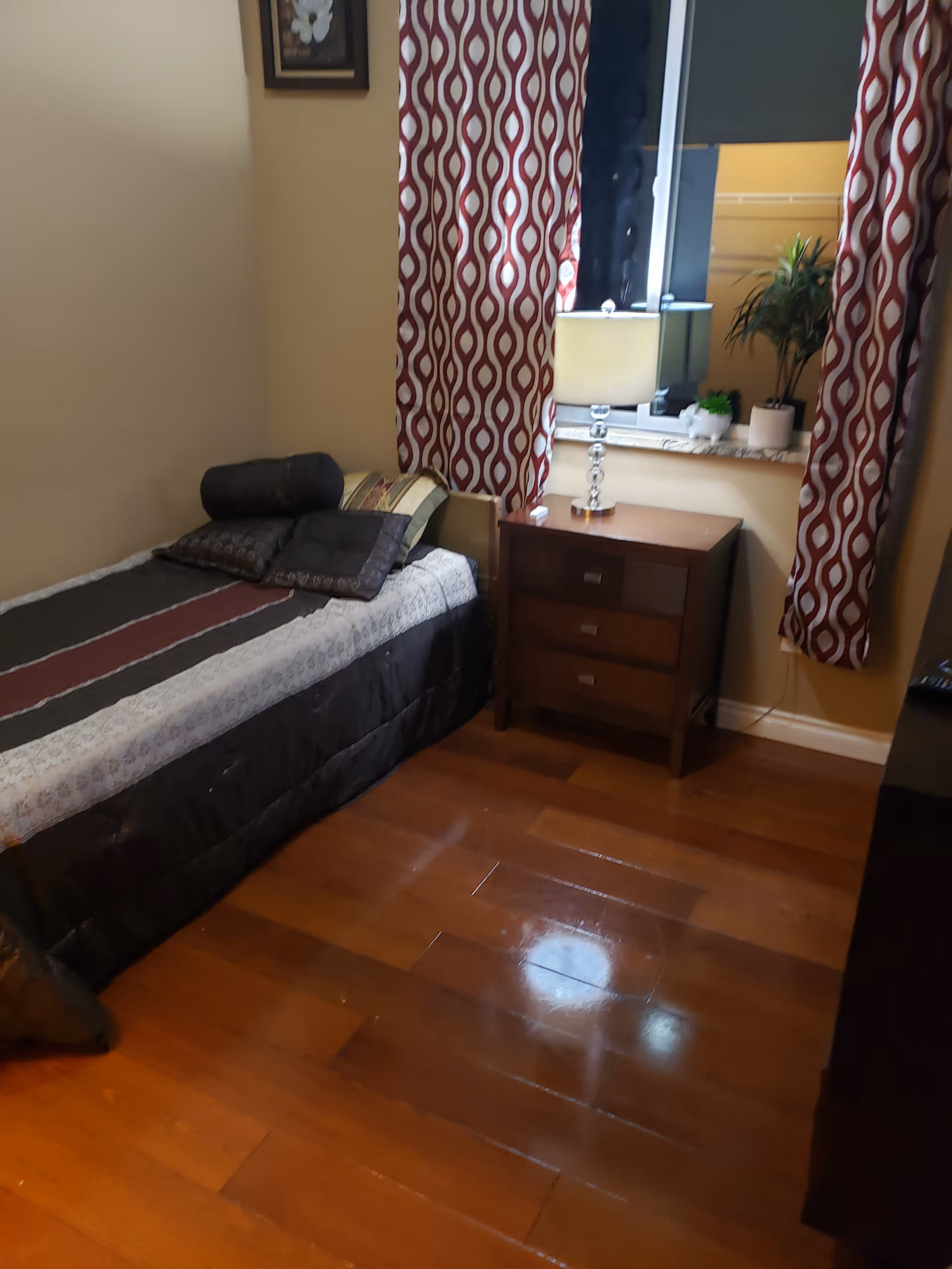 A small bedroom with a single bed covered in a striped bedspread and several pillows. Next to the bed is a wooden nightstand with three drawers, a table lamp, and some small decorative plants on the windowsill. The window is framed by red and white patterned curtains. The floor is made of polished wood.