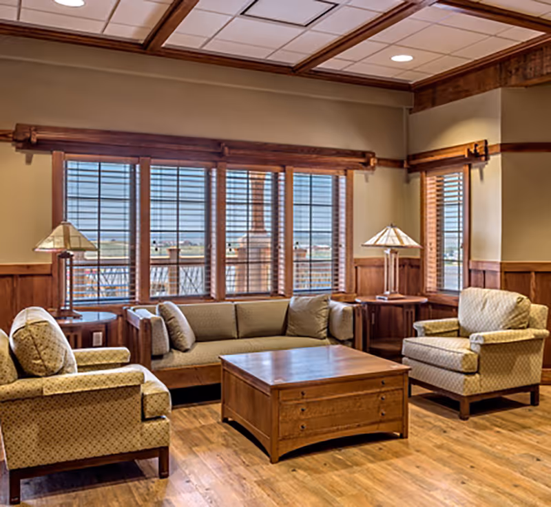 A cozy living room with wooden floors and wood-trimmed walls featuring a beige sofa and two matching armchairs arranged around a wooden coffee table. Two table lamps with stained glass shades sit on side tables next to the sofa and armchairs. Large windows with wooden blinds allow natural light to fill the room.