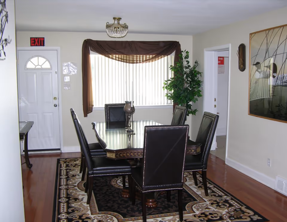 A dining room with a rectangular dark wooden table surrounded by six black leather chairs. The floor has a patterned area rug, and a large window with vertical blinds and a brown valance is on the back wall. There is a white door with a half-circle window on the left side, an exit sign above it, a green potted plant near the window, and a framed artwork on the right wall. The room has light-colored walls and hardwood floors.