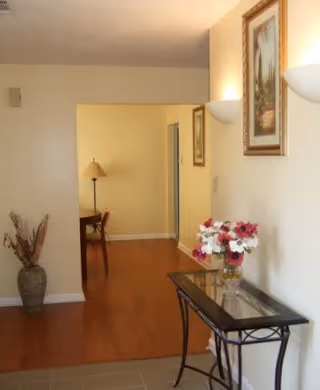 Interior hallway with light-colored walls and wooden flooring leading to a dining area with a table and chairs. A glass-top console table with a vase of flowers is positioned against the right wall, which also has framed artwork and wall-mounted light fixtures. A large decorative vase with dried branches is placed on the left side near the entrance.