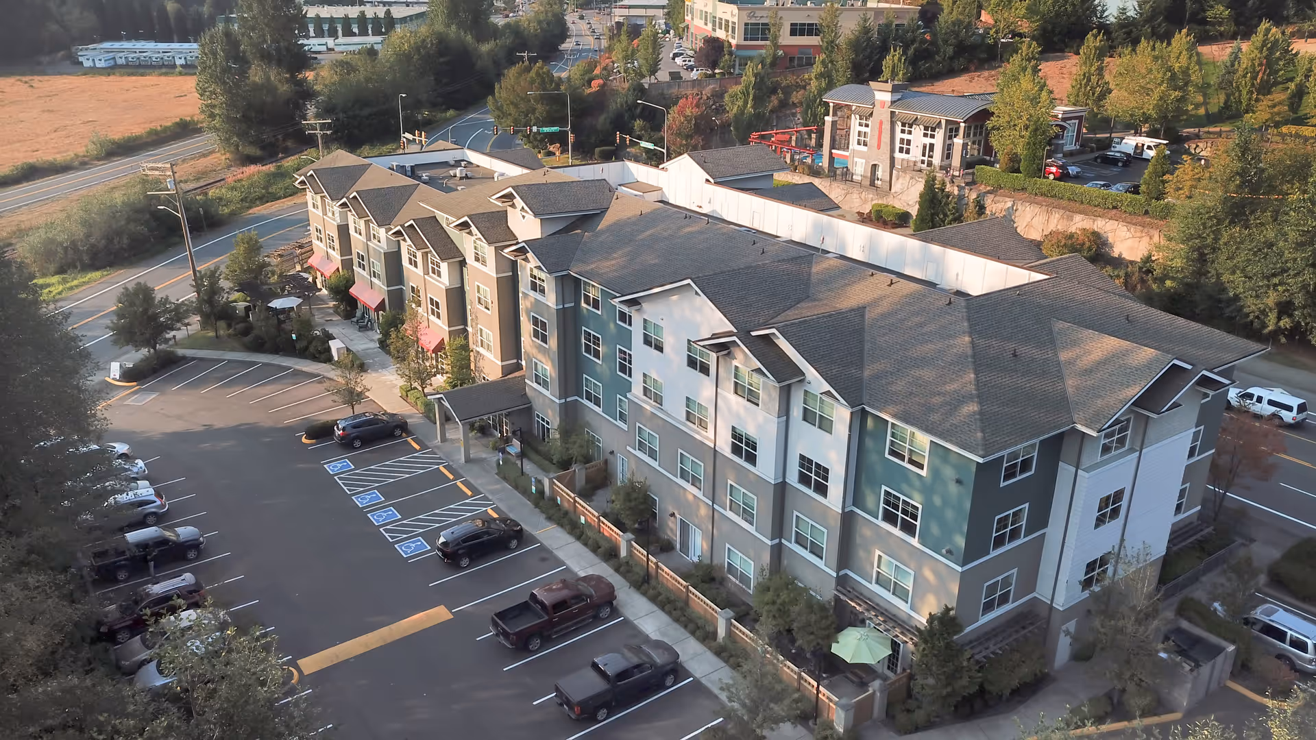 Aerial view of a multi-story senior living facility named The Creekside, showing the building with multiple windows, a parking lot with several cars, and surrounding trees and roads.