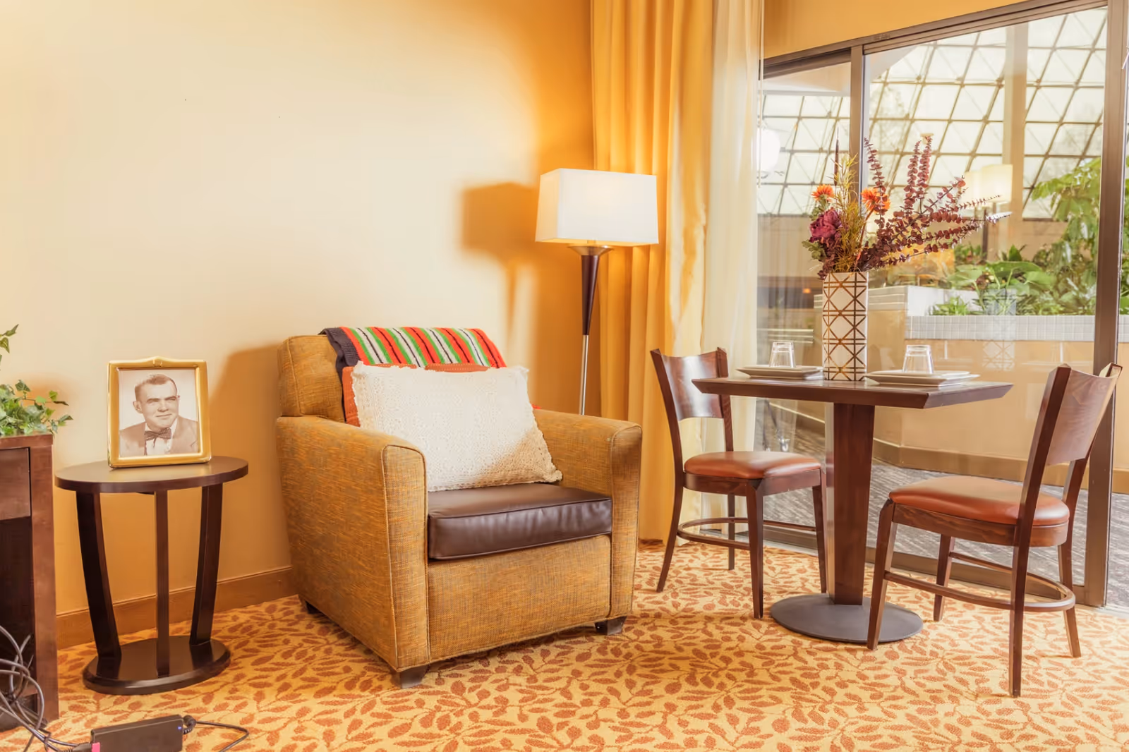Cozy seating area with an armchair, side table holding a framed photo, a floor lamp, and a small round dining table with two chairs by a sliding glass door.