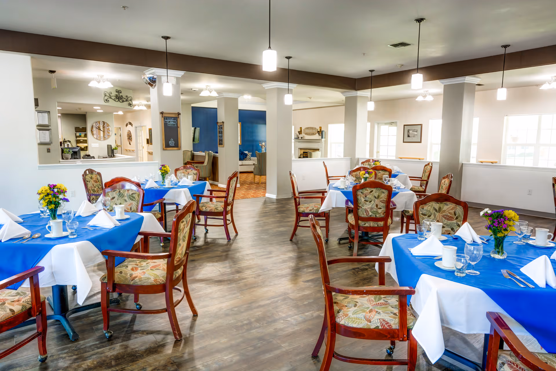 A bright dining room with several tables covered in blue and white tablecloths, each set with white napkins, cups, glasses, and small flower arrangements. The room has wooden chairs with floral upholstery, pendant lights hanging from the ceiling, and a wooden floor. In the background, there are open spaces leading to other rooms with additional seating and decor.