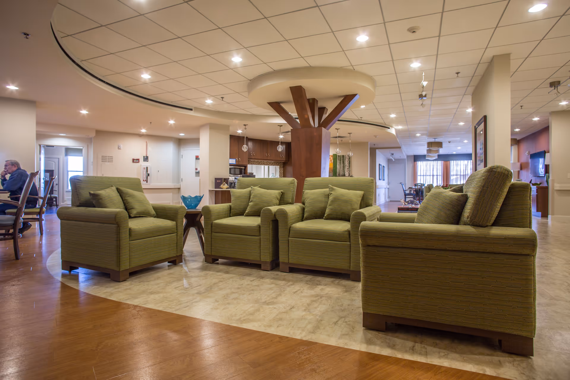 A spacious and well-lit common area in a senior living facility featuring four green upholstered armchairs arranged in a square around a small wooden table with a blue decorative bowl. The room has a mix of wood and tile flooring, a ceiling with recessed lighting, and a central wooden column with a circular ceiling feature. In the background, there are dining tables and chairs, a kitchen area, and large windows with curtains allowing natural light.