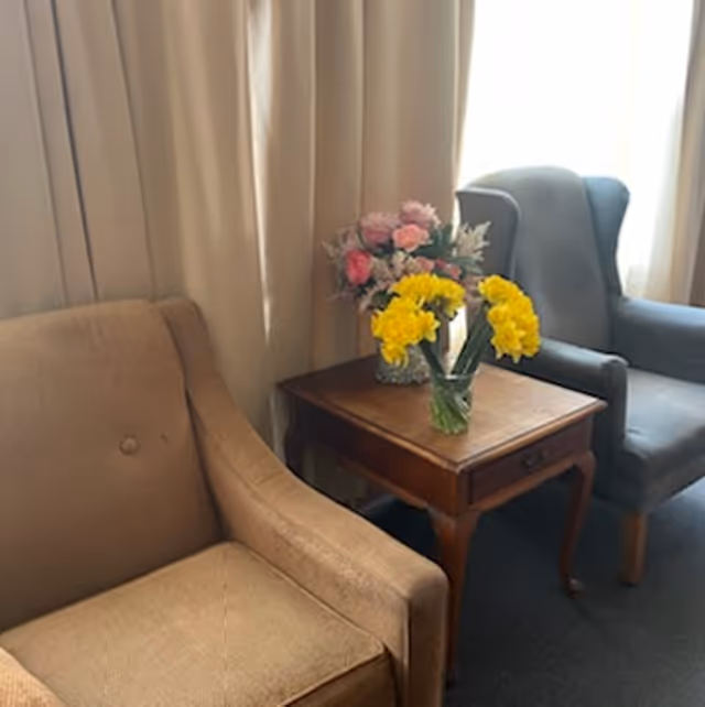A cozy seating area with a beige armchair on the left and a gray armchair on the right, separated by a wooden side table holding two vases of flowers, one with yellow daffodils and the other with pink and white flowers, set against a backdrop of beige curtains.