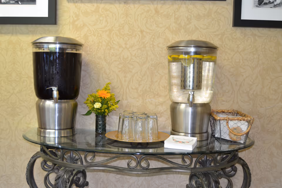 Two beverage dispensers on a glass-topped table with glasses, a small flower arrangement and a basket against patterned wallpaper.