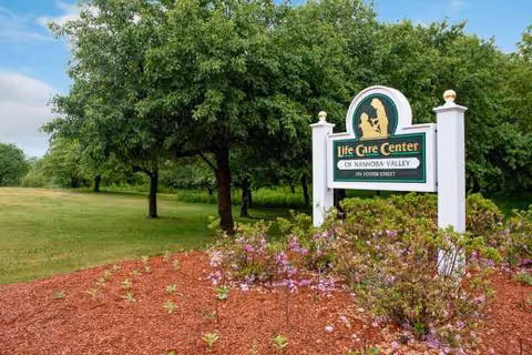 Outdoor view shows the Life Care Center of Nashoba Valley sign with surrounding landscaping.