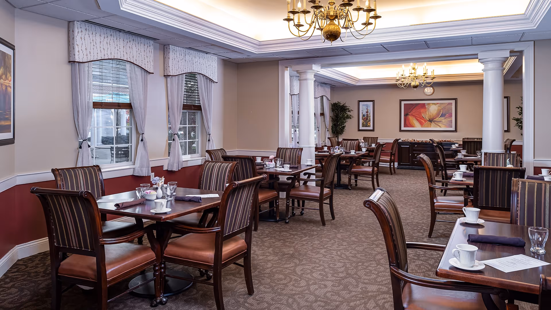 A spacious dining room with multiple wooden tables and chairs arranged neatly. Each table is set with white cups, glasses, napkins, and silverware. The room features large windows with white curtains, beige walls with a red lower half, decorative columns, chandeliers, and framed artwork on the walls.