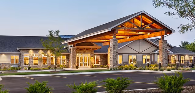 Exterior view of Silverado St. Charles Memory Care Community building at dusk, featuring a large covered entrance with wooden beams and stone pillars, surrounded by landscaped greenery and a parking lot in the foreground.
