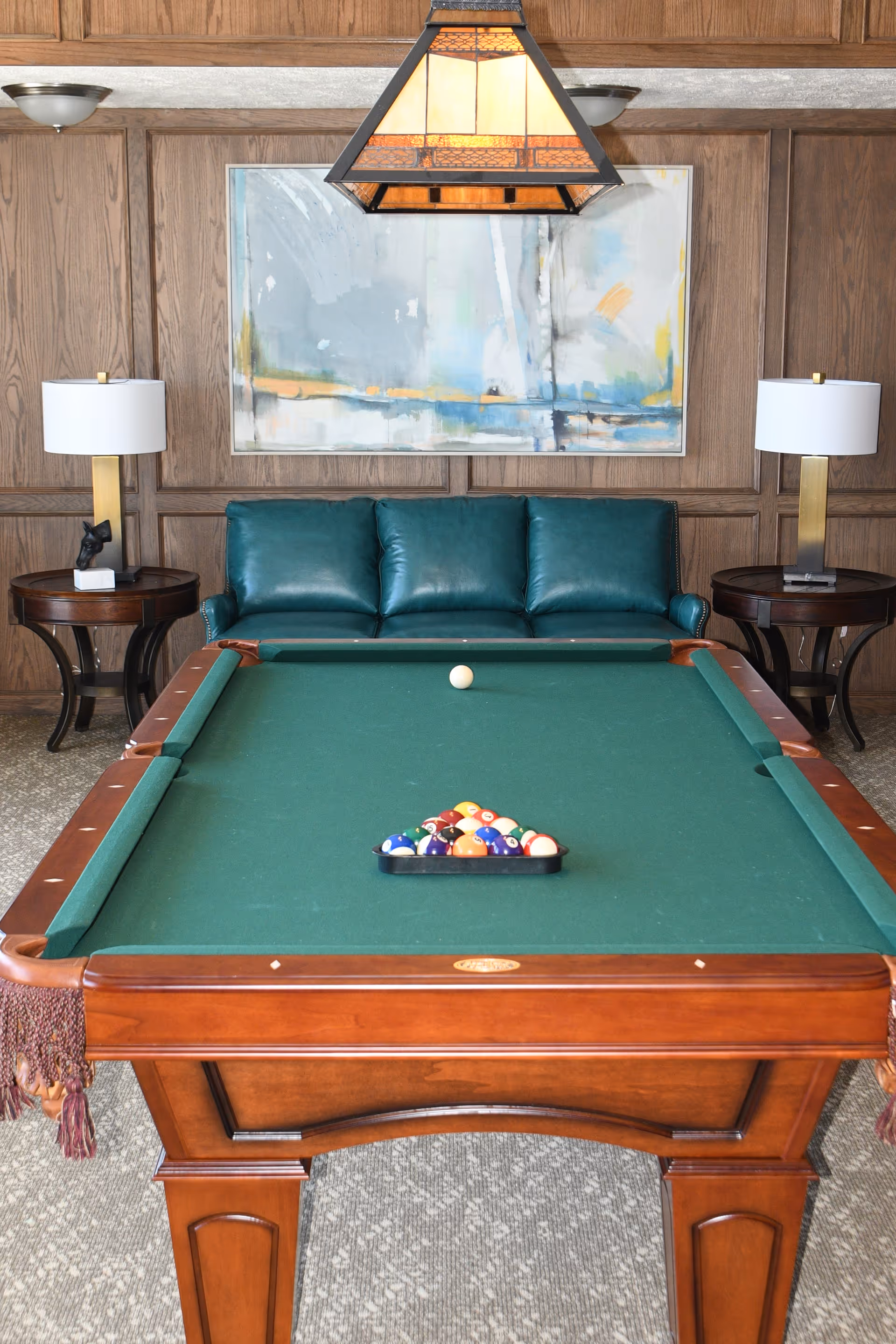 A pool table with a green felt surface and a full set of billiard balls racked in a triangle in the center. Behind the pool table is a teal leather couch flanked by two round wooden side tables, each with a modern lamp. A large abstract painting hangs on the wood-paneled wall, and a stained glass pendant light fixture hangs above the pool table.