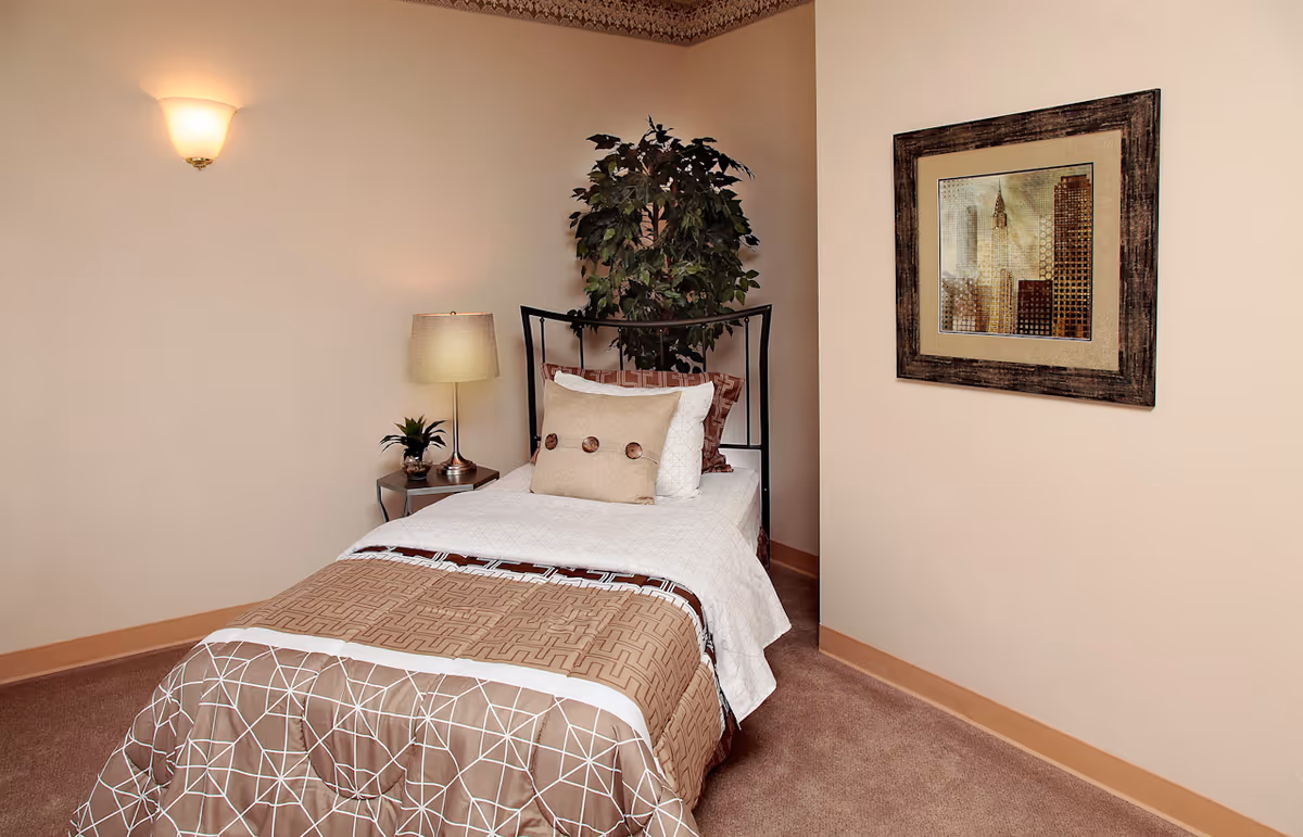 A small bedroom with a single bed covered in white and brown bedding, a side table with a lamp and a small plant, a large potted plant behind the bed, a wall-mounted light fixture, and a framed picture of a cityscape on the wall.