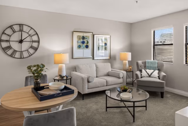 A cozy living room with a light gray loveseat, a gray armchair with a decorative pillow and throw, a round glass coffee table with a small plant, and a round wooden dining table with two gray chairs. The walls are light beige with two framed abstract artworks and a large black metal clock. Two table lamps with beige shades are on side tables next to the loveseat. Two windows with blinds allow natural light into the room.
