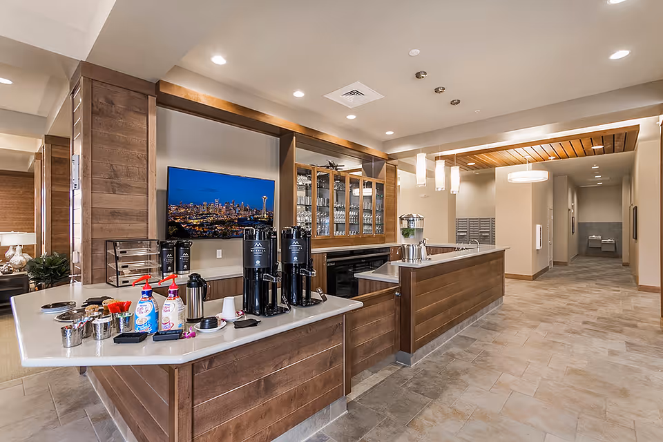 Interior view of a modern common area in a senior living facility featuring a coffee and beverage station with dispensers, cups, and condiments on a countertop. Behind the station is a wall-mounted TV displaying a cityscape at night and glass cabinets with glassware. The area has warm wood paneling, pendant lighting, and a tiled floor leading to a hallway.