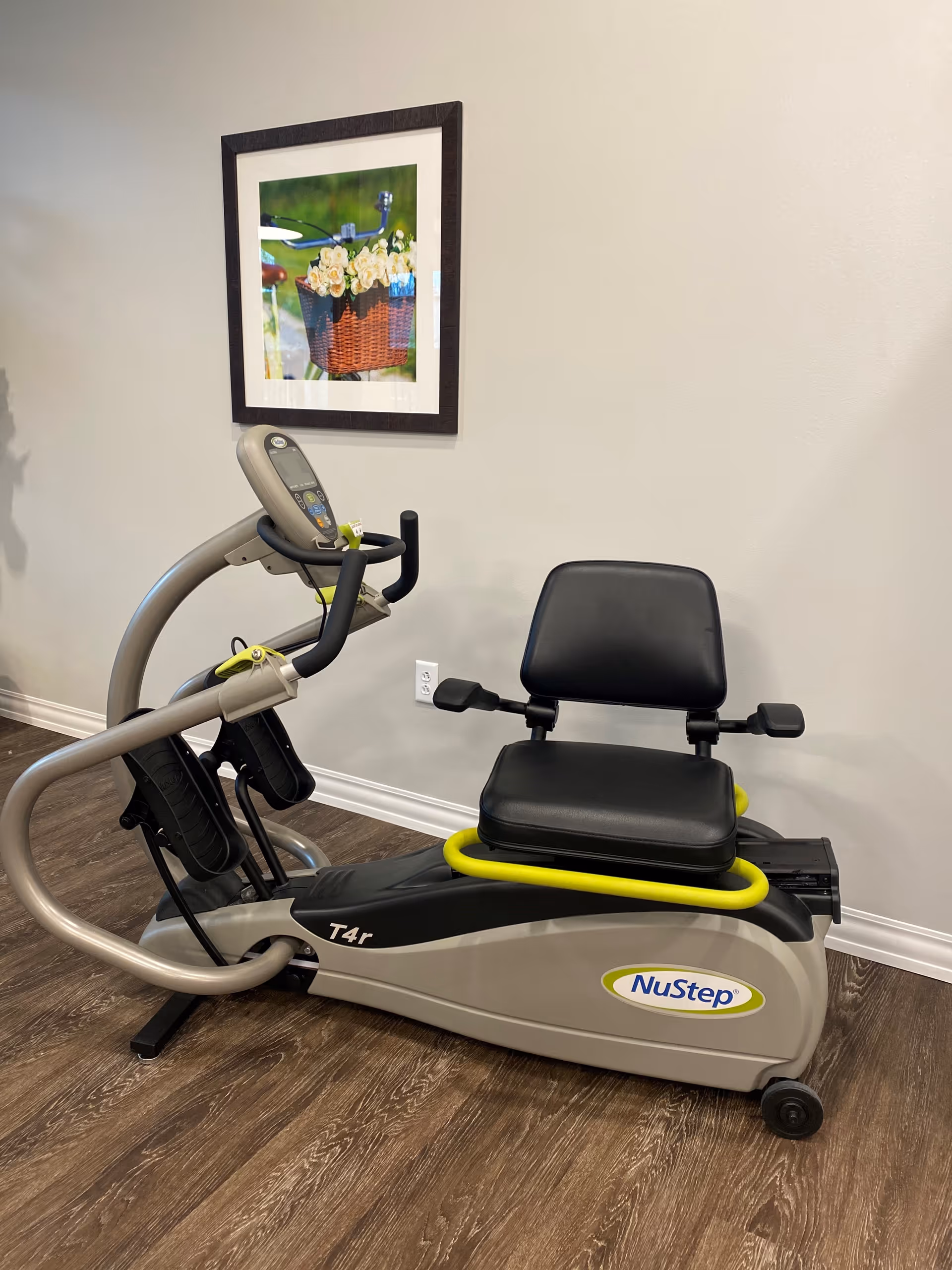 A NuStep T4r recumbent cross trainer exercise machine with a black seat and handles, placed on a wooden floor against a light gray wall. Above the machine, there is a framed picture of a bicycle basket filled with white flowers.