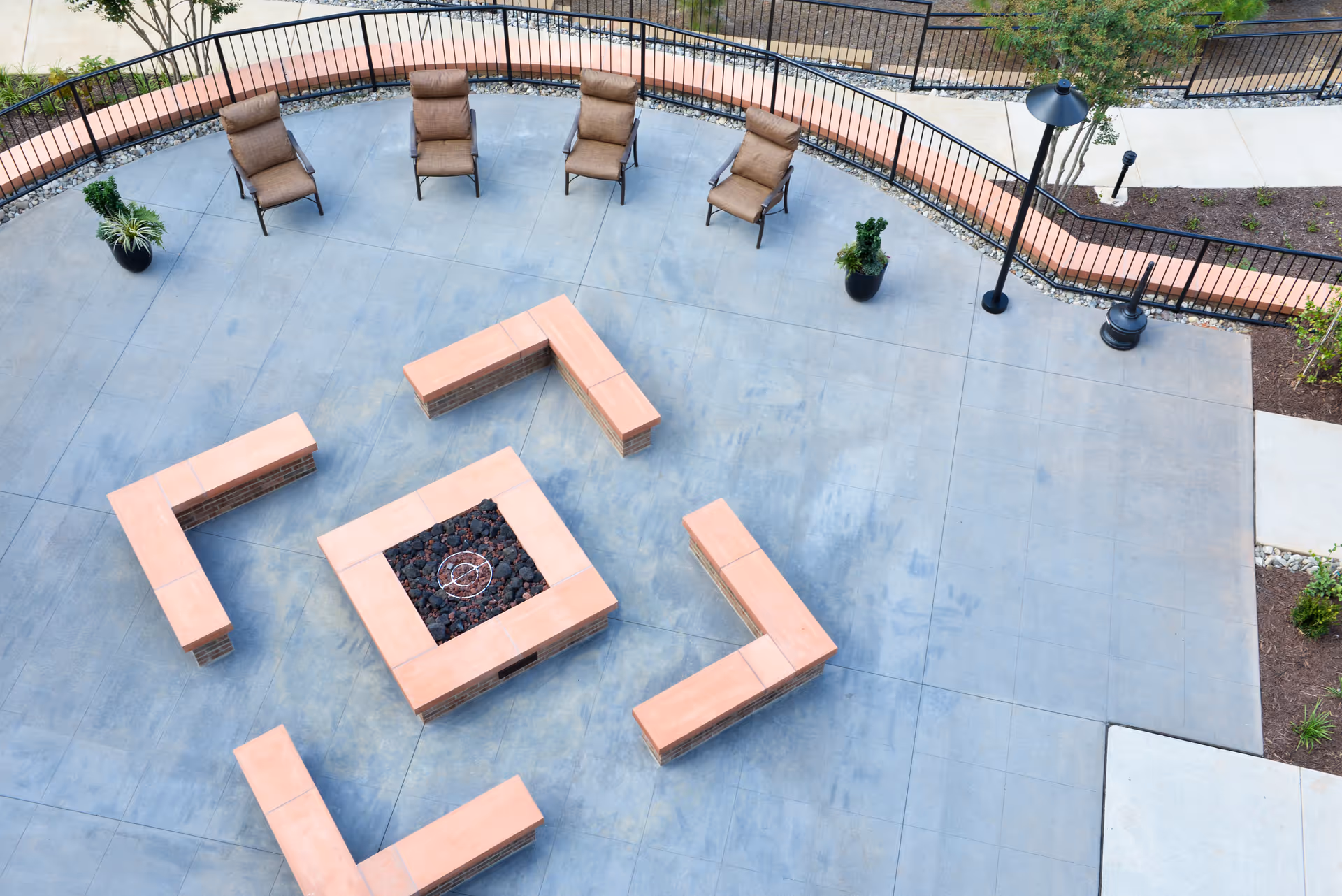 Aerial view of an outdoor patio area with a central square fire pit surrounded by four L-shaped brick benches with terracotta tops. There are five cushioned lounge chairs arranged in a semicircle near a black metal railing. Two potted plants and a black lamp post are also visible on the concrete patio.
