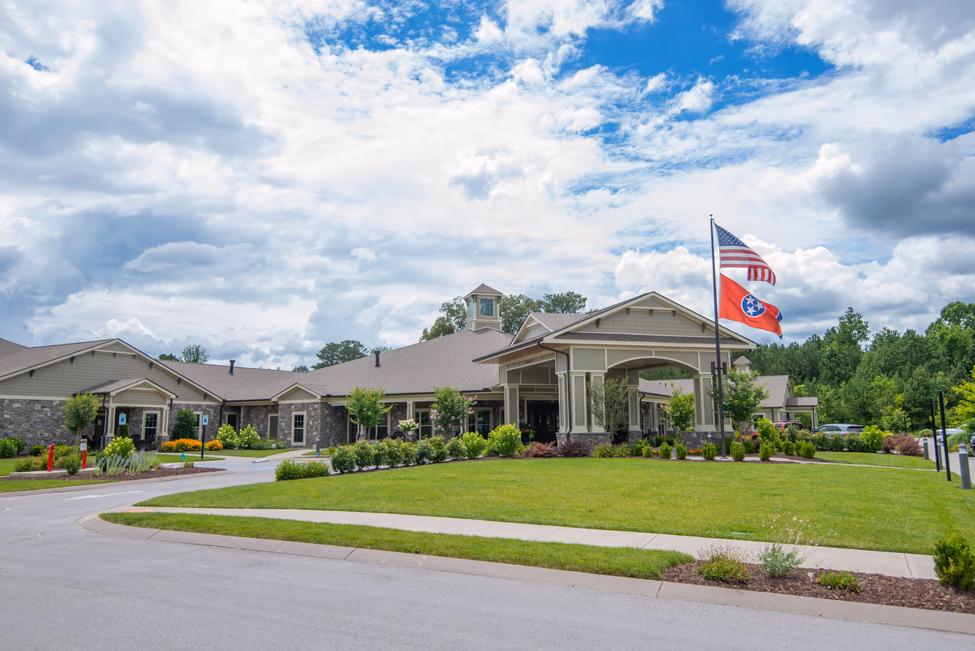 Exterior view of a single-story senior living facility building with a covered entrance, well-maintained lawn, and two flagpoles displaying the American flag and the Tennessee state flag under a partly cloudy sky.
