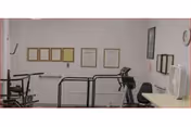 Interior view of a small exercise room with a treadmill, exercise bike, and other fitness equipment. The walls are decorated with framed certificates or documents, and there is a fan on a table to the right.