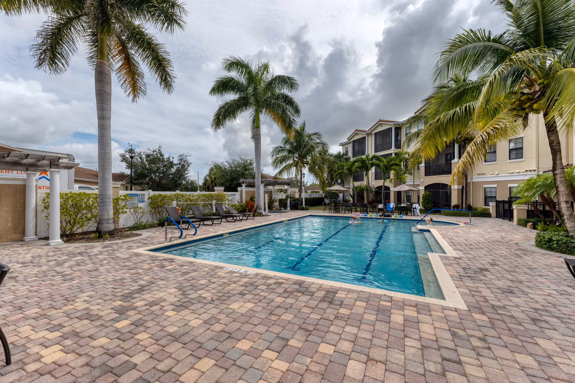 Outdoor swimming pool area at Addington Place of Jupiter with lounge chairs, palm trees, and a multi-story building in the background under a cloudy sky.