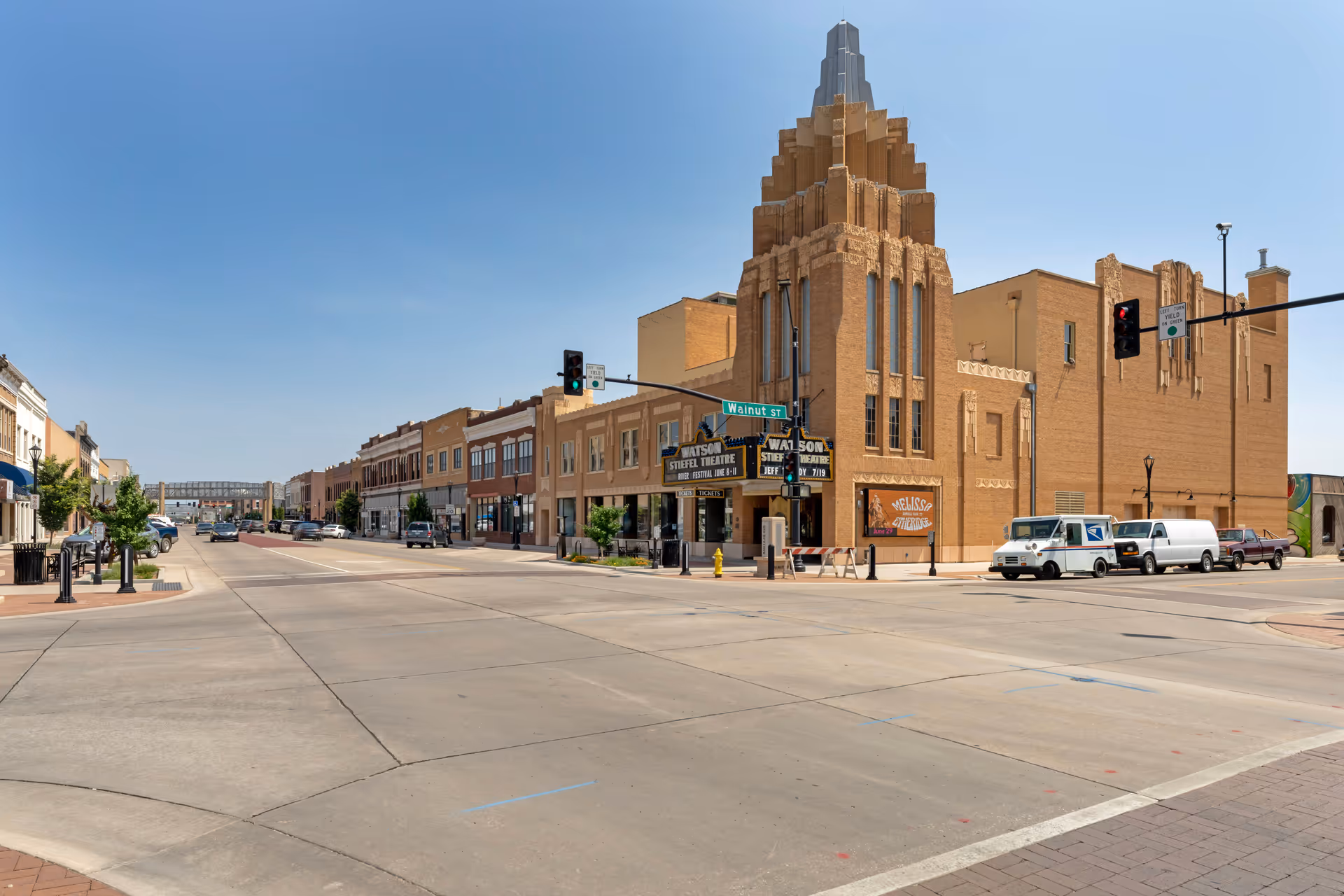 A wide street view of a downtown area with a prominent historic theater building featuring an art deco style tower at the corner. The street is lined with parked cars and other commercial buildings under a clear blue sky.