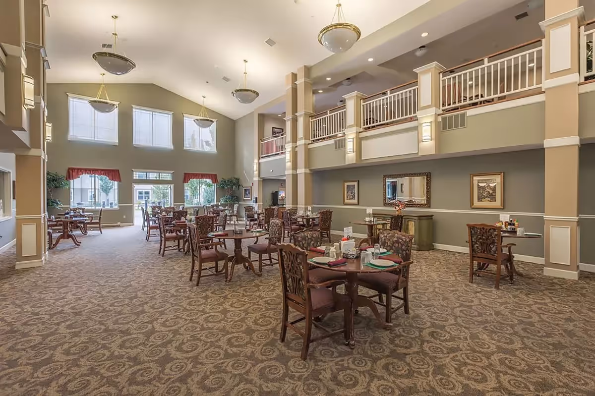 A spacious dining room with multiple round wooden tables and cushioned chairs arranged neatly. The room features high ceilings with large hanging light fixtures, tall windows with red valances allowing natural light to fill the space, and a patterned carpet. There is a second-floor balcony with white railings overlooking the dining area, and framed artwork on the walls.