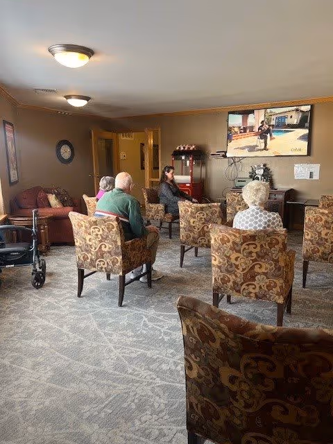 A group of elderly people seated in patterned armchairs in a common room watching a television mounted on the wall. The room has beige walls, carpeted floor, and a few pieces of furniture including a red popcorn machine and a small table with a decorative plant.