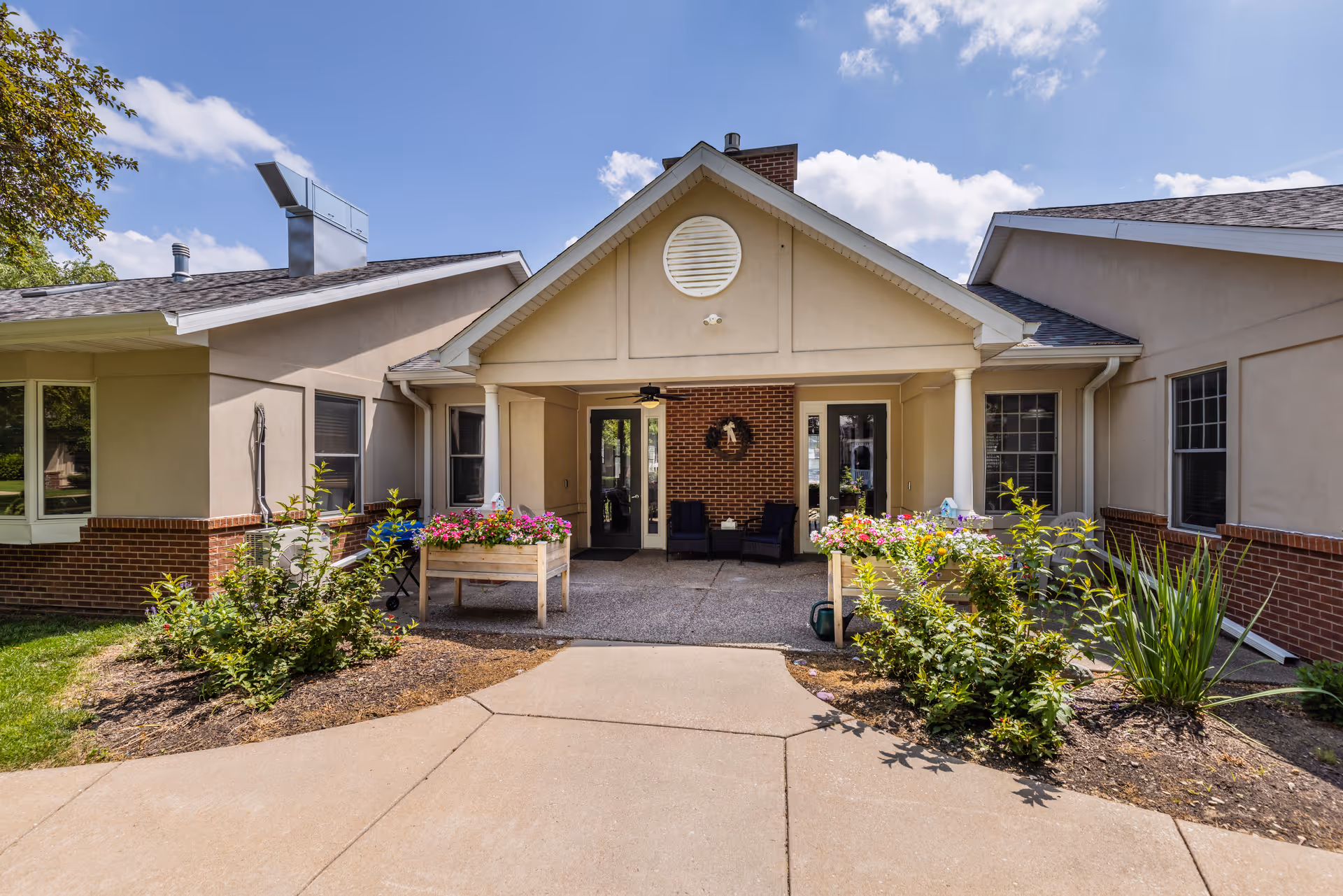 Exterior view of the entrance to a senior living facility with a covered porch area, two planters filled with colorful flowers, and some outdoor seating. The building has beige walls with brick accents and a peaked roof under a partly cloudy sky.