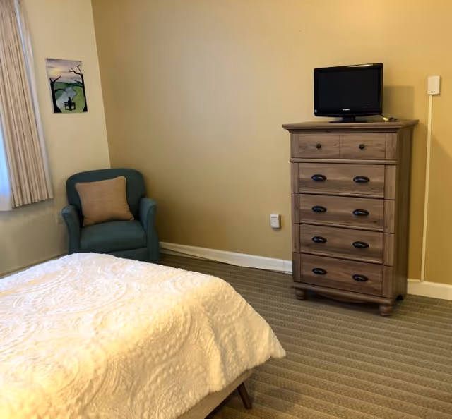 A bedroom with a bed covered in a white textured blanket, a teal armchair with a beige pillow, a wooden dresser with multiple drawers, and a small flat-screen TV on top. The walls are painted beige, and there is a window with beige curtains and a small framed picture on the wall.