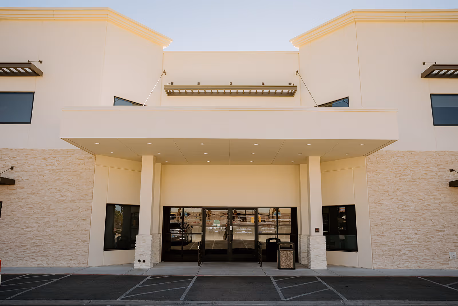 Front exterior view of Green Valley Health & Rehabilitation building with a covered entrance, glass doors, and windows on either side. The building has a beige facade with stone accents and a parking area in front.