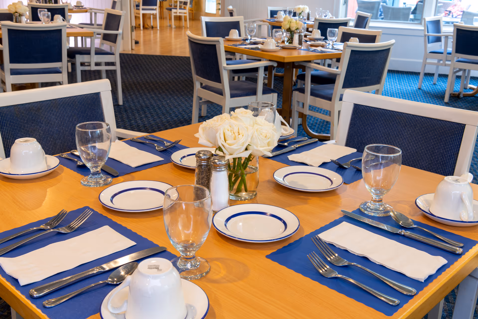 A neatly set dining table with blue placemats, white plates, glassware, silverware, and a vase of white roses in a bright dining room.