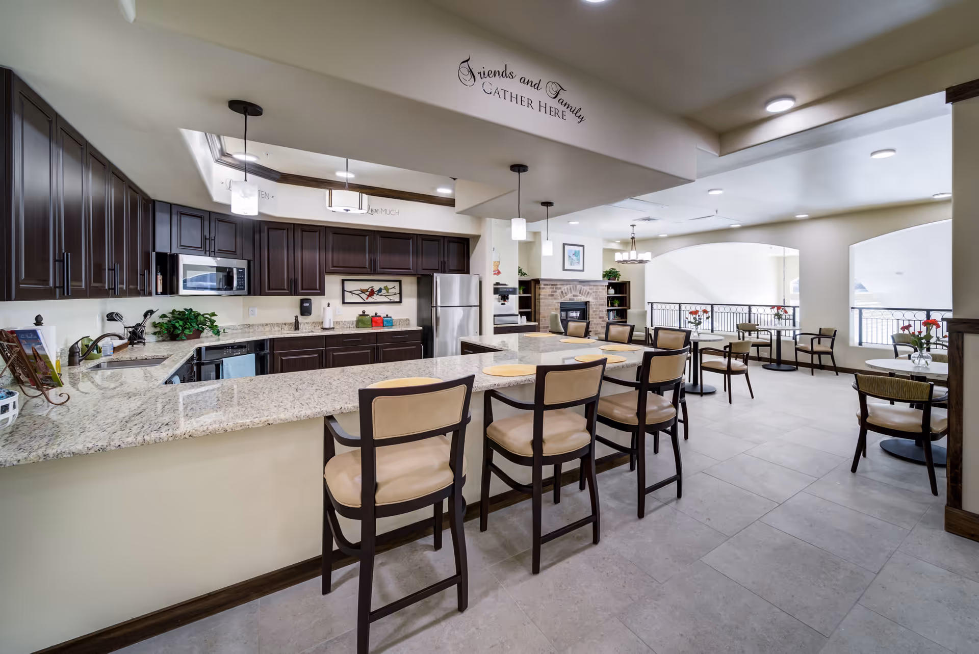 A spacious and modern kitchen and dining area in a retirement living facility. The kitchen features dark wood cabinets, granite countertops, a stainless steel refrigerator, microwave, and dishwasher. There is a large kitchen island with six cushioned bar stools. The dining area has several small round tables with chairs, each table decorated with a vase of red flowers. The walls are light-colored, and the space is well-lit with ceiling lights and pendant lamps. Above the kitchen area, a decorative sign reads 'Friends and Family Gather Here.'