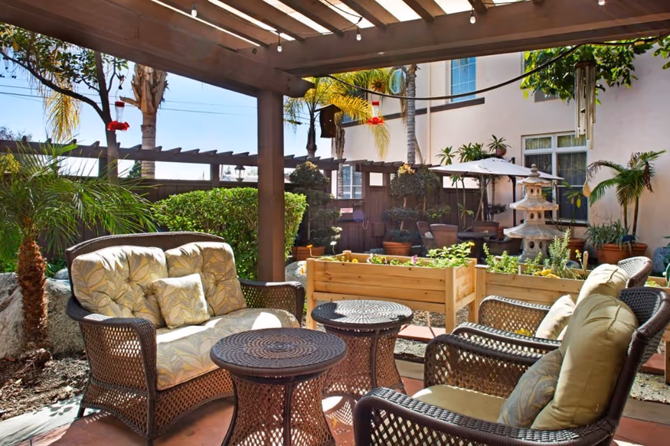 Outdoor patio area with wicker furniture including cushioned chairs and a loveseat under a wooden pergola. There are small round tables, potted plants, raised garden beds, and decorative elements like a stone lantern and hummingbird feeders. The background shows part of a building with windows and an umbrella-covered seating area.