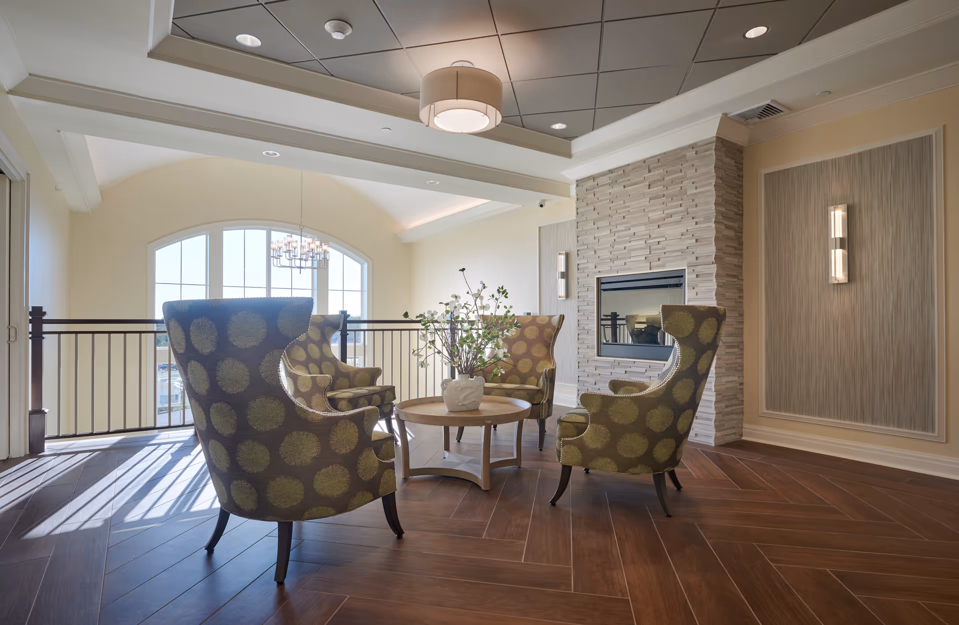 Sunlit lounge with three patterned armchairs arranged around a round table by a fireplace and large arched window.