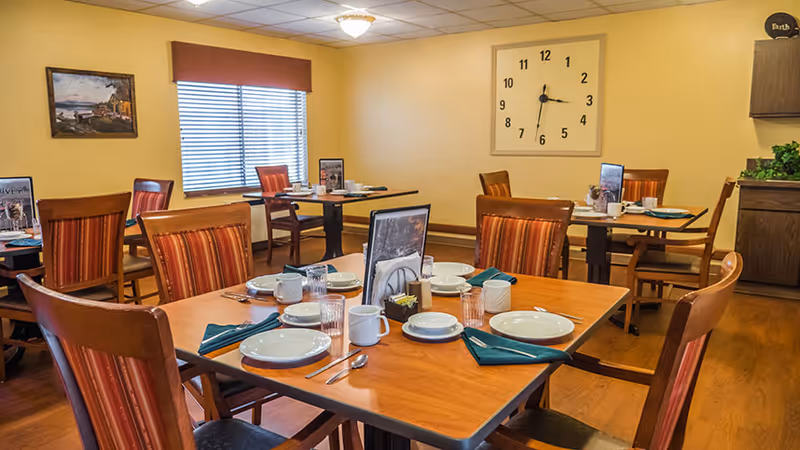 A dining room with wooden tables and chairs set for a meal, including plates, cups, utensils, and green napkins. The room has yellow walls, a large clock on the wall, a window with blinds, and a painting. There is also a cabinet with a plant on top.