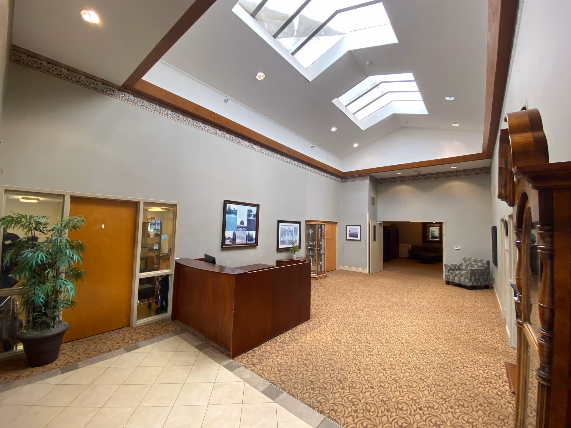 A spacious interior lobby area with a high ceiling featuring skylights. There is a wooden reception desk on the left side, a potted plant next to a wooden door with glass panels, framed pictures on the walls, and a patterned carpet covering the floor. A wooden grandfather clock is visible on the right side, and a cushioned armchair is placed near the back wall.