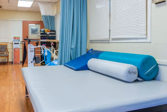 A rehabilitation room with a large padded therapy table covered in a light gray sheet. On the table are blue and teal foam wedges and a cylindrical bolster pillow labeled 'Rehab'. The room has wood flooring, a blue privacy curtain, exercise equipment, a chair, and windows with closed blinds.