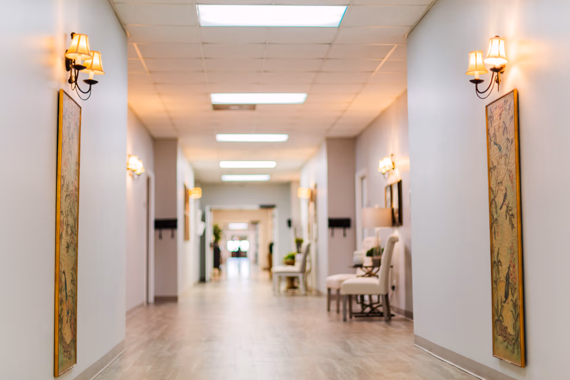 A long, well-lit hallway in a senior living facility with light-colored walls and wooden flooring. The hallway is decorated with wall-mounted lamps and framed artwork. There are chairs and small tables along the walls, and doors leading to rooms on either side.