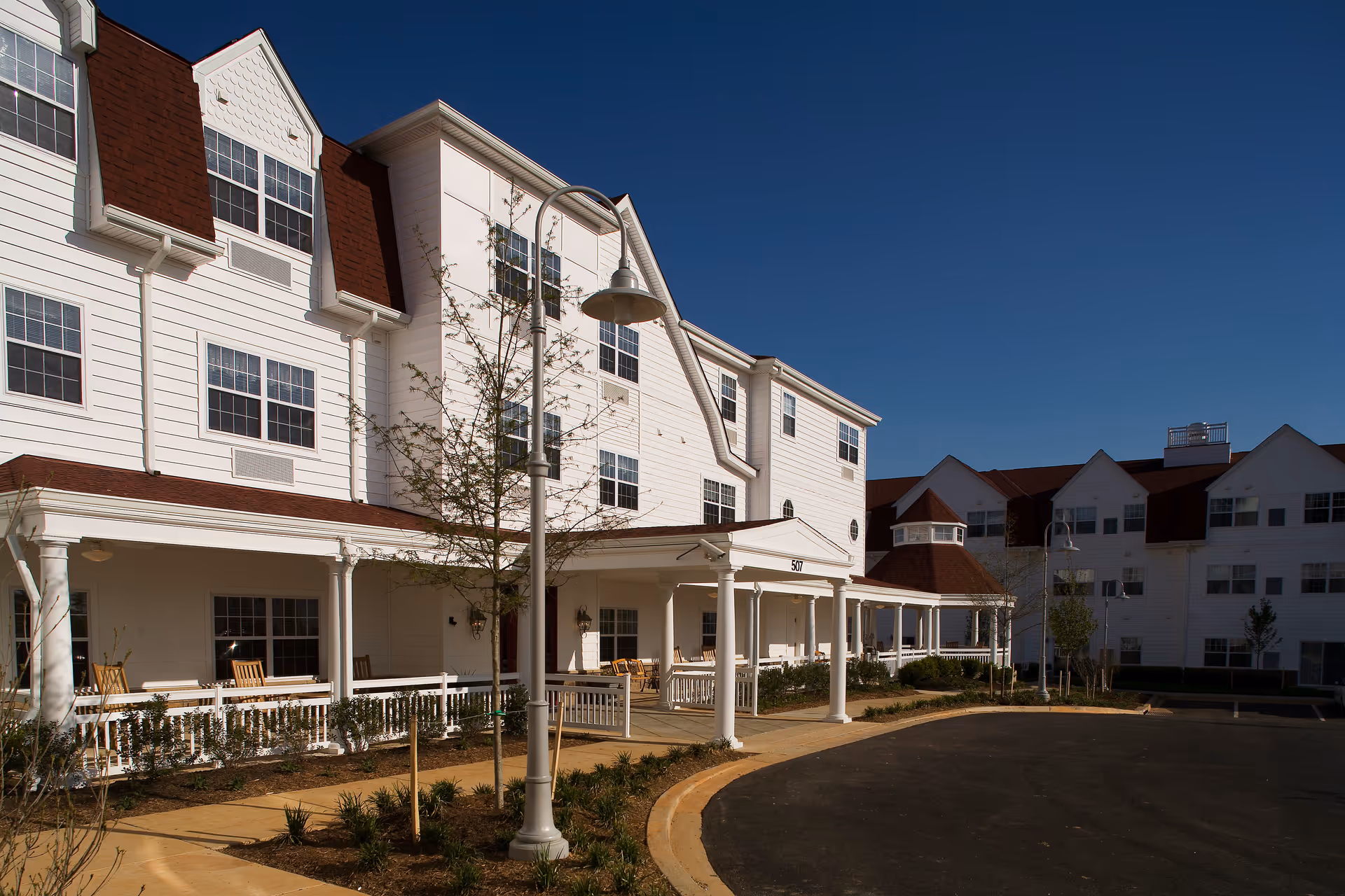 Exterior view of a senior living facility building with white siding and red roofs under a clear blue sky. The building has multiple windows and a covered porch area with rocking chairs. There are young trees and landscaped plants along the walkway and a streetlamp in the foreground.