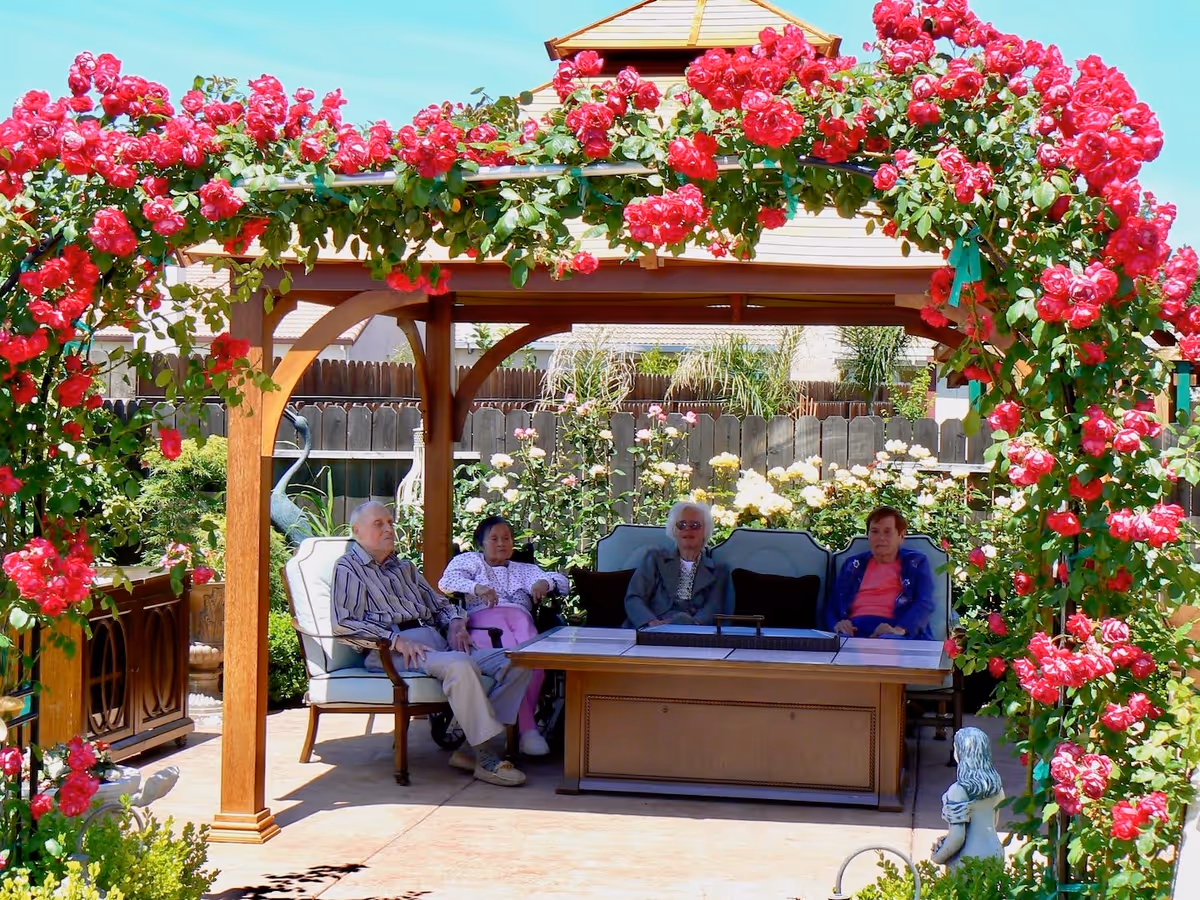 Four elderly people sitting under a wooden gazebo adorned with vibrant red flowers in a garden area, surrounded by greenery and a wooden fence.
