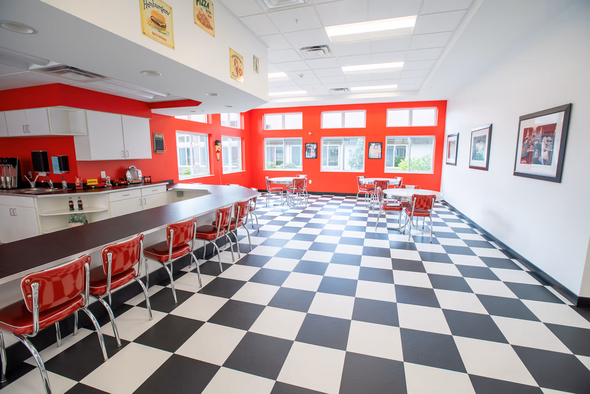 A bright dining area with a retro diner theme featuring black and white checkered flooring, red walls, and red chairs. There is a long counter with red cushioned stools on one side and several small tables with red chairs near large windows letting in natural light. The walls have framed pictures and vintage-style food signs.