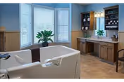 Accessible walk-in bathtub in a bright bathroom with bay windows, plants, and wooden vanities.