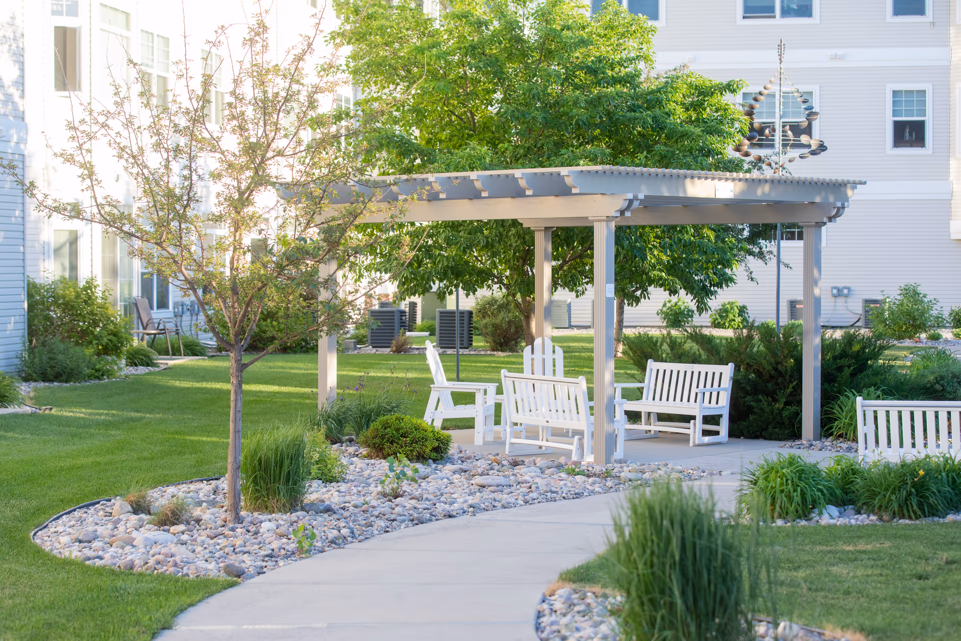 Outdoor seating area in a senior living facility garden with white wooden benches and chairs under a pergola, surrounded by green grass, trees, and landscaping with rocks and plants.