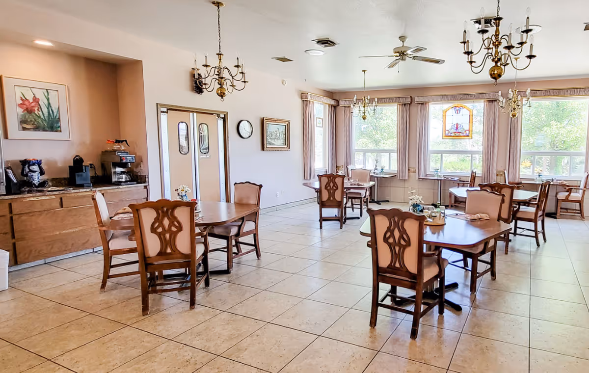 A bright dining room with several wooden tables and chairs arranged neatly. The room features large windows with curtains allowing natural light to fill the space. There are chandeliers hanging from the ceiling and a ceiling fan. On the left side, there is a counter with a coffee machine and some items on it, along with framed artwork on the wall.